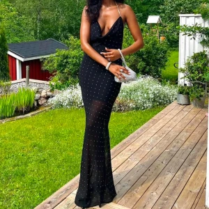 Svart balklänning med glittrigt mönster - Elegant svart långklänning med glittrigt mönster och djup urringning. Klänningen har tunna axelband och en smickrande passform som framhäver figuren. Öppen i ryggen.