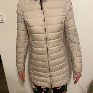 Beige dunjacka från Parajumpers - Säljer en snygg beige dunjacka från Parajumpers. Jackan är superlätt och har en dragkedja framtill. Den har en huva och långa ärmar, perfekt för kyliga dagar. Märkeslogga på ärmen ger en extra touch. Passar perfekt för vintern! ❄️
