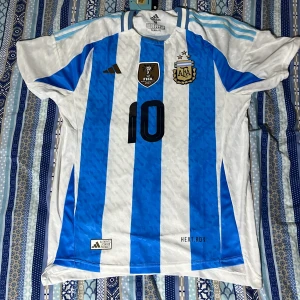 Argentina fotbollströja med nummer 10 - Säljer en officiell Argentina fotbollströja från Adidas med nummer 10 och namn på ryggen. Tröjan är vit och blå med randigt mönster och har korta ärmar. Den har FIFA World Champions 2022-märke och AFA-logga på bröstet. Är helt nytt och oanvänd!