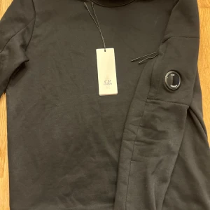 C.P. Company tröja - Säljer en stilren svart långärmad tröja från C.P. Company. Tröjan har en unik detalj på ärmen och är perfekt för en modern look. Märkets logga finns både på etiketten och som en snygg detalj. Köpt på CareOfCarl, kvitto finns. Nypris 2199kr