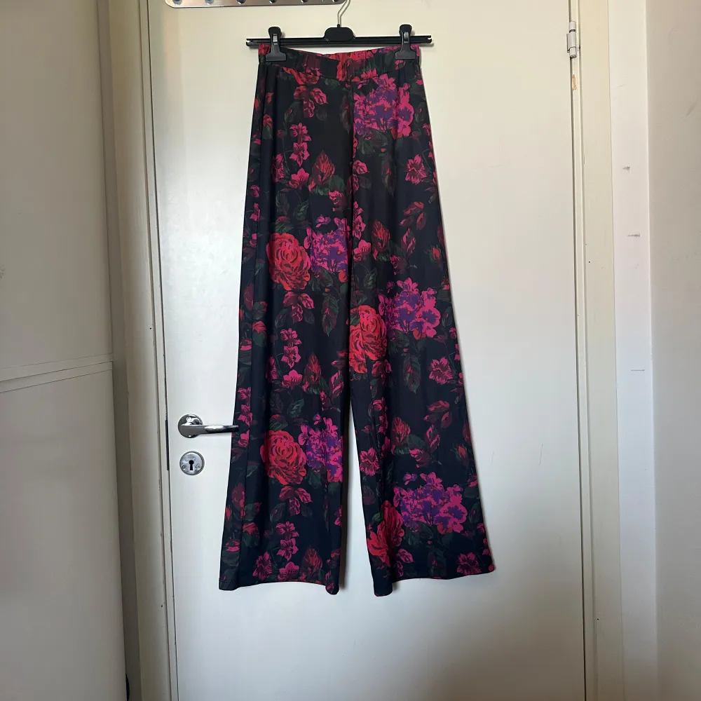 Snygga svarta byxor med ett färgglatt blommönster i rött och rosa. Byxorna har en loose passform och är perfekta för att ge en färgklick till din outfit. De är högmidjade och har en rak stil som ger en avslappnad look. Storlek XS. Men de är stora i storlek. Mycket fint skick. . Farkut & Housut.