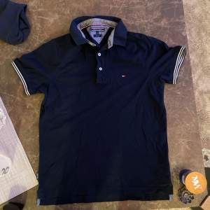 Snygg mörkblå pikétröja från Tommy Hilfiger med klassisk krage och knappar framtill. Tröjan har korta ärmar med randiga detaljer i vitt och blått. Perfekt för en stilren look.