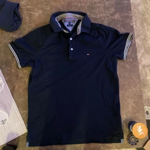 Mörkblå pikétröja från Tommy Hilfiger - Snygg mörkblå pikétröja från Tommy Hilfiger med klassisk krage och knappar framtill. Tröjan har korta ärmar med randiga detaljer i vitt och blått. Perfekt för en stilren look.