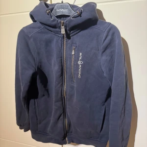 Sail Racing zip hoodie - Säljer en blå fleecejacka från Sail Racing med dragkedja och huva. Hoodien är i storleken 150 och är i helt okej skick och är ganska så använd och sen är det någon fläck på den som borde gå bort i tvätten.