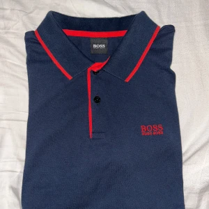 Mörkblå pikétröja från Hugo Boss - Snygg mörkblå pikétröja från Hugo Boss med röda detaljer. Tröjan har en klassisk krage och korta ärmar. Perfekt för en stilren look.