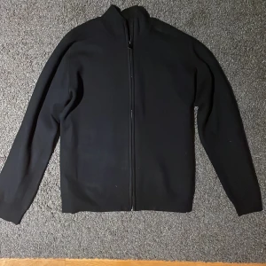 Stickad zip up - Snygg svart stickad tröja med dragkedja och hög krage. Perfekt för en stilren look. Tröjan har långa ärmar och är tillverkad i ett mjukt material som ger en bekväm passform. Helt ny
