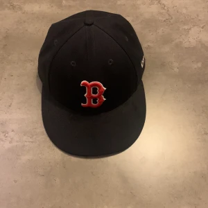 Mörkblå La keps  - Snygg mörkblå keps från New Era med ett broderat rött 'B' framtill. Perfekt för sportfantaster och vardagsbruk. Kepsen har en klassisk design och är tillverkad i bomull för en bekväm passform.