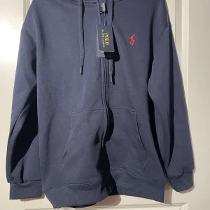 Mörkblå hoodie från Ralph Lauren - Säljer en snygg mörkblå hoodie från Ralph Lauren med dragkedja och broderad logga i rött på bröstet. Tröjan har långa ärmar och en praktisk huva med snörning. Perfekt för en avslappnad stil.