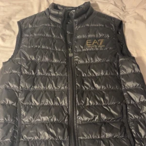 Svart dunväst från Emporio Armani - Säljer en stilren svart dunväst från Emporio Armani. Västen har en dragkedja framtill och är quiltad för extra värme. Perfekt för kyligare dagar och ger en snygg look med sin diskreta design.