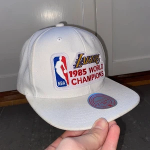 Vit keps med Lakers broderi - Snygg vit keps från Mitchell & Ness med Lakers 1985 World Champions broderi. Kepsen har en platt skärm med grön undersida och justerbar passform. Perfekt för basketfans!