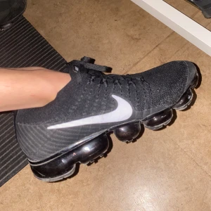 Nike Vapormax  - Säljer ett par svarta Nike Vapormax med vit swoosh. Skorna har en unik sula med synliga luftkuddar för extra komfort och stil. Perfekta för både träning och vardag. Skorna har snörning för en säker passform. Den har inga fel ingenting behöver bara torkas av med en ren trasa så ser den helt ny ut. Nypris 2100kr. Öppen för byten och kan diskutera om priset