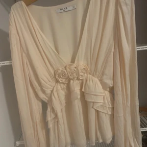 Beige blus från NA-KD - Säljer en elegant beige blus från NA-KD med v-ringning och långa ärmar. Blusen har vackra volangdetaljer och tre dekorativa rosor framtill.