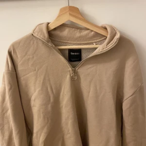 Beige quarter zip från Bershka - Säljer en beige zip från Bershka med dragkedja och mysigt foder. Perfekt för kyliga dagar med sin bekväma passform och stilrena design. Passar till många olika outfits!