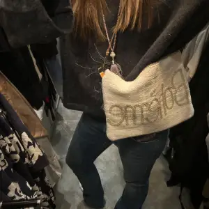 Trendig beige axelväska med broderad text 'Boheme' på framsidan. Väskan har en justerbar rem i läder och en praktisk dragkedja upptill. Perfekt för att ge en bohemisk touch till din outfit.