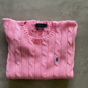 Rosa stickad tröja från Ralph Lauren - Säljer en snygg rosa stickad tröja från Ralph Lauren i Pima Cotton. Tröjan har ett klassiskt kabelstickat mönster och en broderad logga i blått på bröstet. Perfekt för en stilren look! Aldrig använd