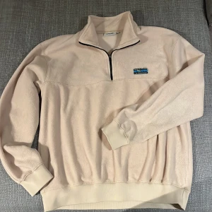 Beige fleece tröja från Junkyard  - Säljer en beige fleece tröja från Junkyard med halv dragkedja och broderad logga på bröstet. Tröjan har långa ärmar och ribbade muddar vid ärmslut och nederkant. Perfekt för kyliga dagar!