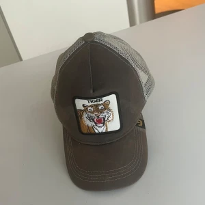 Brun keps med tigerbroderi - Snygg brun keps med ett coolt tigerbroderi framtill. Kepsen har ett nätmaterial baktill. Storleken justerar man genom flärpen där bak. Den är använd men är fortfarande i ett bra skick. 🙌