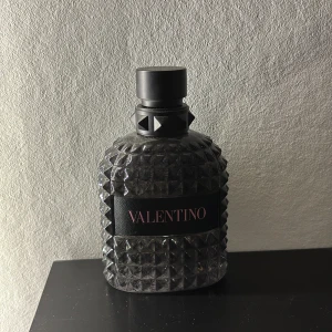 Valentino Uomo 100ml - Snygg parfymflaska från Valentino med en unik design. Flaskan är täckt av pyramidformade nitar och har en elegant svart etikett med märkesnamnet i rött. Den svarta korken matchar flaskans stilrena utseende. Perfekt för den som vill ha en doft med stil och attityd.