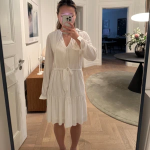 Vit ribbad klänning med knytband - Säljer en stilren vit klänning med ribbad struktur och v-ringning. Klänningen har långa ärmar och ett knytband i midjan för en justerbar passform. Perfekt för en elegant look. 