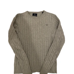 Grå Gant tröja - Säljer en stilren grå stickad tröja från GANT i stretchig bomull. Tröjan har ett diskret märke på bröstet och ribbade muddar vid ärmslut och nederkant. Perfekt för en avslappnad och snygg look.