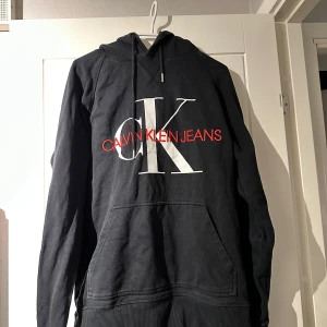 Svart hoodie från Calvin Klein - Säljer en stilren svart hoodie från Calvin Klein med stor vit CK-logga och röd text på framsidan. Tröjan har en klassisk känguruficka och justerbar huva med snören. Perfekt för en avslappnad look. Skriv för fler frågor. Pris kan diskuteras vid snabb affäf🤩