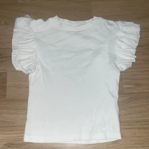 Vit topp med volangärmar från Gina Tricot - Säljer en superfin vit topp från Gina Tricot i storlek 146/152. Den har korta ärmar med volangdetaljer som ger en söt och trendig look. Perfekt för både vardag och fest!. passar en tajt xs också 