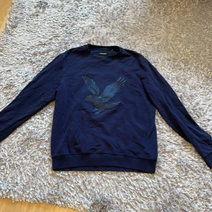 Mörkblå tröja från Lyle & Scott - Säljer en snygg mörkblå tröja från Lyle & Scott med ett coolt örntryck på framsidan. Tröjan är långärmad och perfekt för både höst och vinter. Den är i bra skick.