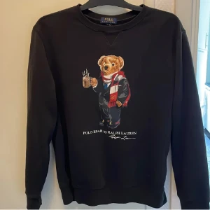 Svart tröja med Polo Bear från Ralph Lauren - Säljer en svart tröja från Ralph Lauren med den ikoniska Polo Bear på framsidan. Björnen har en röd och vit halsduk och håller en varm dryck. Tröjan är perfekt för kyliga dagar och har en klassisk passform. Den är i bra skick och redo för en ny ägare! skriv vid minsta tanke + pris kan diskuteras vid snabb affär!