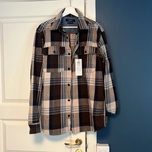Rutig skjorta från Jack & Jones - Säljer en snygg rutig skjorta från Jack & Jones i bruna, beige och blå nyanser. Skjortan har långa ärmar och knappar framtill, perfekt för en avslappnad stil. Materialet känns mjukt och bekvämt, vilket gör den idealisk för både vardag och fest.