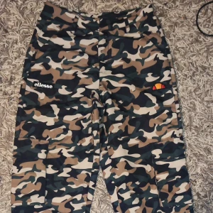 Camouflage cargobyxor från Ellesse - Säljer ett par coola camouflage cargobyxor från Ellesse. De är perfekta för en avslappnad stil och har praktiska fickor på sidorna. Byxorna är i en snygg mix av gröna, bruna och beige toner. Perfekta för höst och vår, och de har en normal passform. Perfekt för dig som vill sticka ut lite!