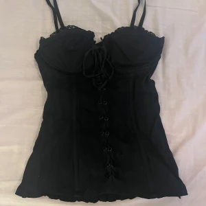 Svart korsett med spetsdetaljer - Snygg svart korsett med spetsdetaljer och snörning framtill. Den har justerbara axelband. skriv för frågor