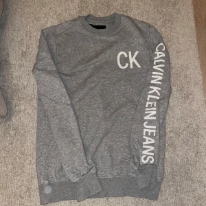 Grå sweatshirt från Calvin Klein Jeans - Säljer en snygg grå sweatshirt från Calvin Klein Jeans i storlek XS. Tröjan har långa ärmar och ett tryck med 'CK' på bröstet och 'CALVIN KLEIN JEANS' längs ena ärmen. Skriv vid minsta lilla fundering✍️😀✅