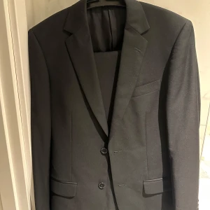 Svart kavaj - Snygg svart kavaj i mycket bra skick. Perfekt för både fest och formella tillfällen. Kavajen har en klassisk design med två knappar och fickor fram. Materialet är en blandning av polyester och viskos, vilket ger en skön passform. Storleken är 50 i kavajen och byxorna är 30-32, vilket motsvarar herr M. Passar utmärkt till höst och vinter.