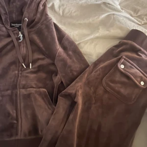Brun velourträningsdräkt från Juicy Couture - Säljer en supermjuk och bekväm brun velourträningsdräkt från Juicy Couture. Hoodien har dragkedja och snörning, medan byxorna har en praktisk ficka med knappdetalj. Perfekt för en avslappnad dag eller mysiga stunder hemma. 🧸har likadan i grå skriv för intresse säljer för 800🫰🏽