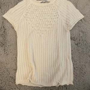 Vit stickad topp från Zara - Säljer en söt vit stickad topp från Zara i storlek S. Den har ett fint mönster framtill och korta ärmar. Perfekt för vår och sommar! 🌼