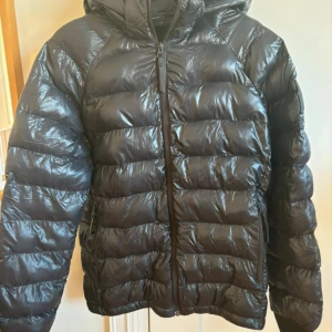 Svart dunjacka från Peak Performance - Säljer en snygg och varm svart dunjacka från Peak Performance. Jackan har en glansig finish och är perfekt för kalla dagar. Den har en dragkedja framtill och en mysig huva. Perfekt för vintern! Har dock hål i fickan men inget som syns utifrån! Skriv om ni har några funderingar💕