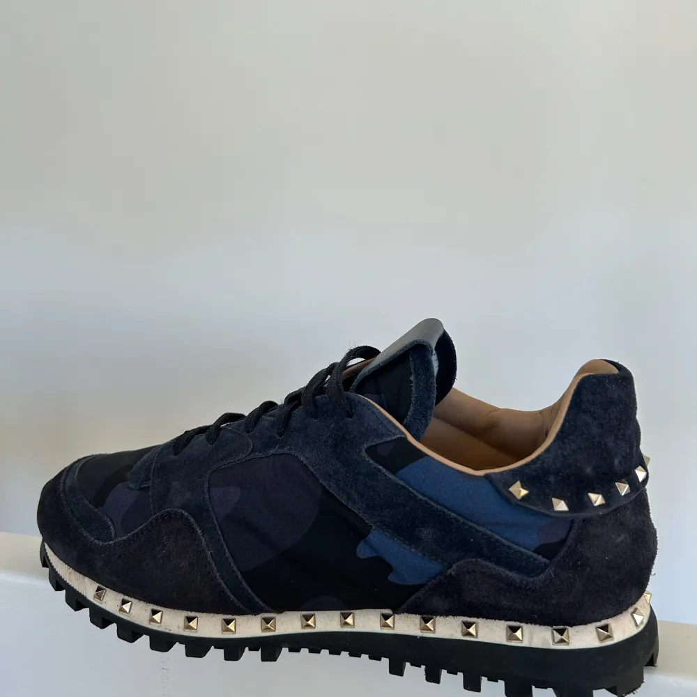 Snygga Valentino Garavani Rockrunner sneakers i mörkblå mocka med coola nitar runt sulan. De har ett diskret camouflage-mönster och klassisk snörning. Perfekta för att ge din outfit en lyxig touch. Skorna är i mycket bra skick och har inga synliga defekter! . Skor.