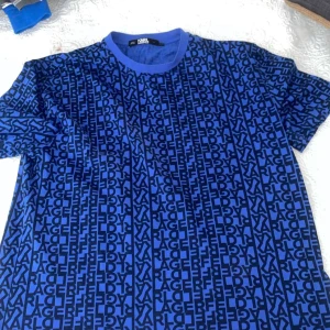 Blå t-shirt från Karl Lagerfeld - Snygg blå t-shirt från Karl Lagerfeld med ett coolt mönster av svarta bokstäver över hela tyget. Den är kortärmad och perfekt för en avslappnad stil. Passar bra till både jeans och shorts. Perfekt för sommarens alla äventyr! 
