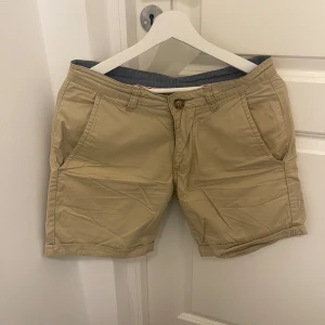 Beige shorts från Lager 157 - Säljer ett par snygga beige shorts från Lager 157 i storlek M. De är gjorda av 100% bomull och har en klassisk design med knappar och bälteshällor. Perfekta för sommardagar eller en avslappnad vårlook. Shortsens stilrena design gör dem lätta att matcha med olika outfits.