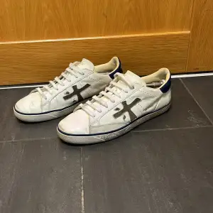 Säljer ett par vita sneakers från Premiata i bra skick. Skorna har en cool grå detalj på sidan och mörkblå detaljer vid hälen. De är perfekta för vardagsbruk och har en klassisk snörning. Sulan är svart och ger bra grepp. Passar perfekt för både höst och vinter 