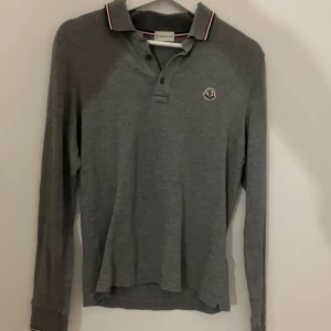 Grå långärmad pikétröja från Moncler - Säljer en stilren grå långärmad pikétröja från Moncler. Den har en klassisk krage med röd och vit randdetalj och knappar framtill. Perfekt för både vardag och lite finare tillfällen. Tröjan är i bra skick och passar perfekt till höst och vår.
