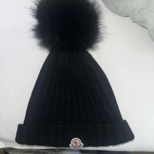 Svart stickad mössa från Moncler - Snygg svart stickad mössa från Moncler med en stor fluffig pälsboll på toppen. Den har en uppvikt kant och Monclers logga framtill. Perfekt för att  hålla sig varm och stilren under vintern. Nypris 3250 kr jag säljer den för 1000 kr. Pris går och diskutera 