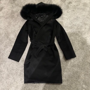 Svart kappa från Hollies - Säljer en elegant svart kappa från Hollies i storlek 36. Den har en lyxig äkta päls runt huvan och ett knytbälte i midjan för en snygg passform. Perfekt för kyliga höst- och vinterdagar. Kappan är i mycket bra skick. Säljer den då jag köpte den här på Plick men den var för liten! 