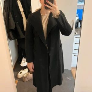 Svart kappa med knytband - Säljer en elegant svart kappa . Den har långa ärmar och ett snyggt knytband i midjan för en figursydd look. Perfekt för höst, vår eller vinter! 🖤Har mite noppor men är fortfarande väldigt fin