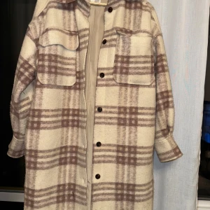 Rutig lång kappa - Säljer en superfin lång kappa i beige och brunt rutigt mönster. Den har långa ärmar och knappar framtill. Perfekt för höst och vår, och ger en stilren look. Passar bra till både vardag och fest. Lite oversize
