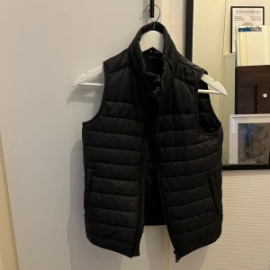 Svart dunväst - Säljer en stilren svart dunväst som är perfekt för kyliga dagar. Den ger en snygg look över en tröja eller hoodie. Passar både för vardag och lite mer uppklädda tillfällen. Västen har storlek 146/152 men passar som en XS