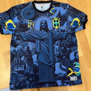 Blå fotbollströja med Brasilienmotiv - Säljer en unik blå fotbollströja med ett stort tryck av Kristusstatyn i Rio de Janeiro. Tröjan har korta ärmar och detaljer i gult och grönt som representerar Brasiliens flagga. Perfekt för fotbollsfans som vill sticka ut!