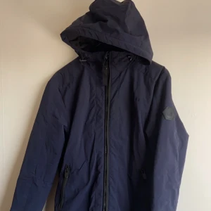 Vinterjacka  - Säljer en snygg mörkblå jacka från FSBN. Den har en dragkedja framtill och en praktisk huva. Perfekt för vinter och hlst med sin stilrena design. Jackan har långa ärmar och ribbade muddar vid ärmslut och nederkant. Märkeslogga på ärmen ger en extra touch. Passar både till vardags och lite finare tillfällen.