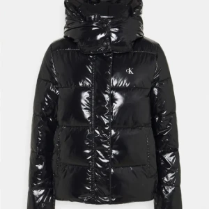 Svart glansig pufferjacka från Calvin Klein - Säljer en snygg svart pufferjacka från Calvin Klein i glansigt material. Jackan har en hög krage och en stor huva för extra värme. Perfekt för kalla vinterdagar! Den har en dragkedja framtill och CK-loggan på framsidan. Frakt ingår. 