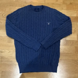 Blå kabelstickad tröja från GANT - Säljer en snygg blå kabelstickad tröja från GANT i premium bomull. Perfekt för kyliga dagar med sin klassiska design och ribbade muddar. Tröjan har en liten broderad logga på bröstet. Mycket fint skick. Passar både till vardags och lite finare tillfällen. 🧶💙